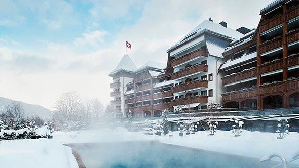 Зимний фасад отеля The Alpina Gstaad с подогреваемым открытым бассейном на фоне заснеженных гор