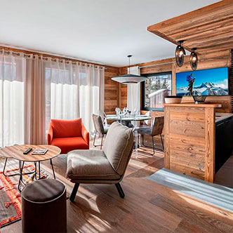 Светлая открытая гостиная-столовая апартаментов Le Blanchot в Вал-д&rsquo;Изер: оранжевое кресло, прозрачные стулья, панорамные окна с видом на снежные склоны.