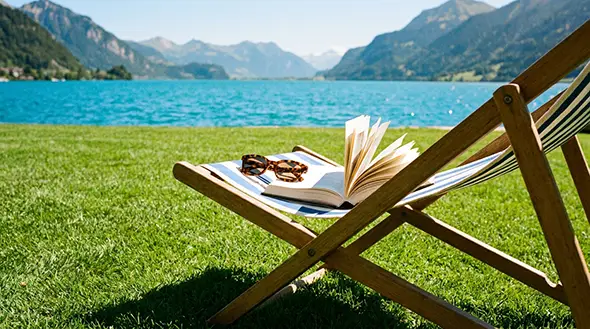 Striped deck chair with a book on the green shore of a clear lake.