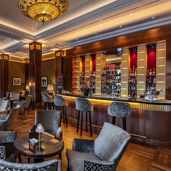 Роскошный интерьер Anton&rsquo;s Bar с деревянными панелями, мягкими креслами с узором, длинной барной стойкой с золотистыми стеллажами и массивной люстрой. / Luxurious interior of Anton&rsquo;s Bar featuring wood paneling, patterned upholstered armchairs, a long bar counter with golden shelving, and a massive chandelier.