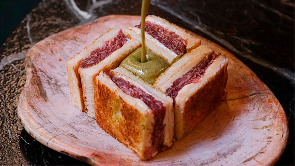 Премиальный Croque Sando с начинкой из мраморной говядины (Wagyu) в поджаренном тостовом хлебе, поливаемый зеленым соусом, на деревянной тарелке
