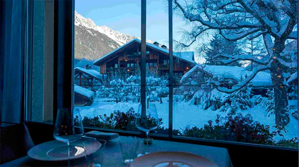 Вид на заснеженное шале и горы из панорамного окна ресторана в отеле Hameau Albert 1er в Шамони