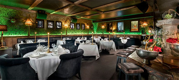 Изысканный интерьер La Mangeoire с ужином, переходящим в вечеринку: столы накрыты белыми скатертями, бархатные стулья, стены украшены рельефными зелеными панелями, картинами и золотистым декором.