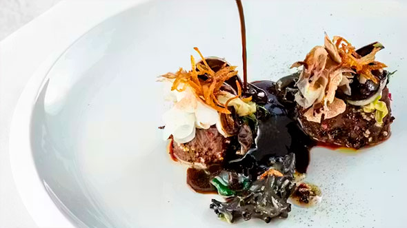 Процесс подачи блюда: рука шефа поливает темным насыщенным соусом два порционных рулета из мяса/овощей, украшенных жареным луком или грибами, на белоснежной тарелке