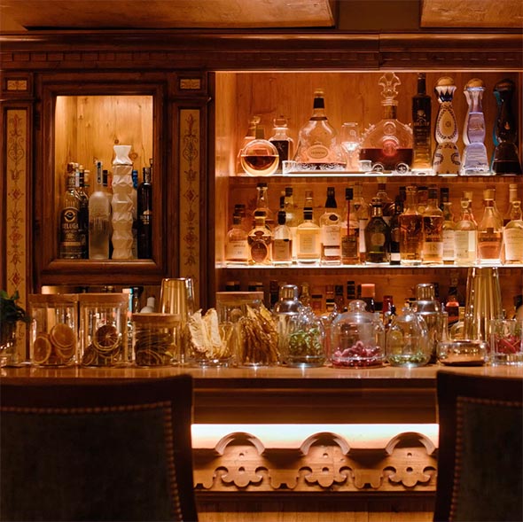 Крупный план элегантной деревянной барной стойки Bar / Tea Room Airelles с подсветкой, где выставлены бутылки премиального алкоголя на полках и декоративные банки с ингредиентами для коктейлей (сушеные фрукты, ягоды) на переднем плане.