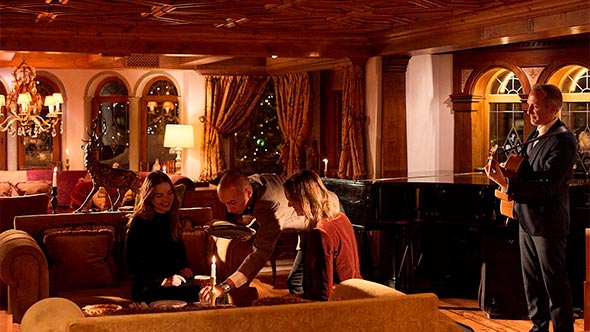 Уютный лаундж Bar / Tea Room Airelles: гости сидят на роскошных диванах с подушками, обитыми бархатом, в центре интерьера, украшенного деревянными панелями и потолком с резьбой; справа стоит рояль и певец в костюме с гитарой.