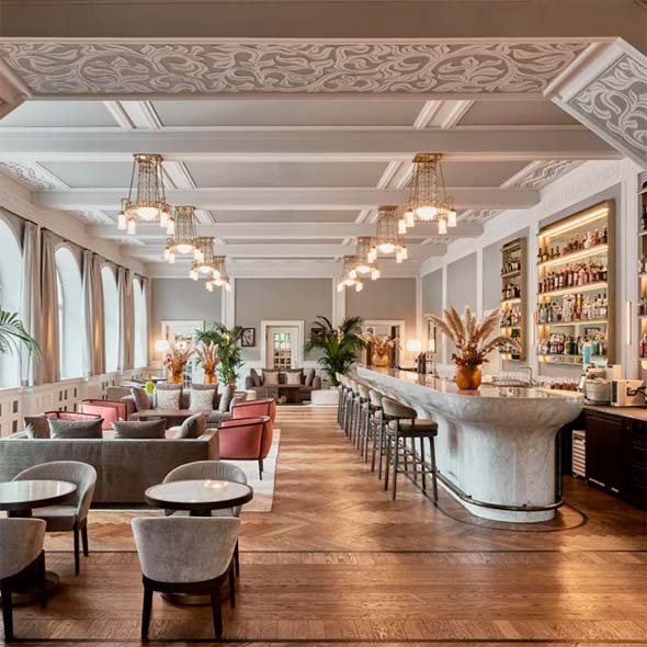 Стильный и светлый интерьер N/5 The Bar с высоким потолком, декоративной лепниной, длинной барной стойкой из белого мрамора и лаунж-зоной с мягкой мебелью. / Stylish and bright interior of N/5 The Bar featuring a high ceiling, decorative stucco, a long white marble bar counter, and a lounge area with soft seating.