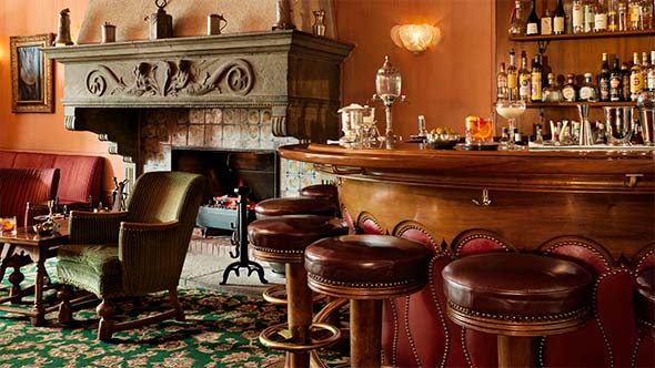 Уютный уголок лаунж-бара с обитыми тканью креслами (зеленым и красным) у массивного каменного камина, над которым скрещены две сабли для фехтования. / Cozy corner of a lounge bar with upholstered armchairs (green and red) by a massive stone fireplace, above which two fencing swords are crossed.