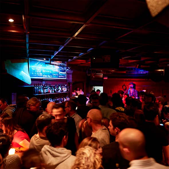 Переполненный людьми бар St&uuml;bli Bar с приглушенным красным освещением, где у дальней стены стоит барная стойка и играет живая музыка. / Crowded St&uuml;bli Bar interior with dim red lighting, featuring a bar counter against the far wall and live music being played.