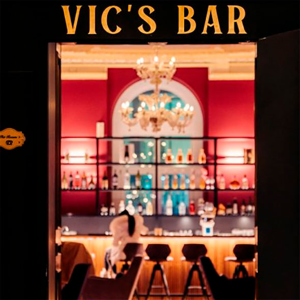 Вход в бар Vic&rsquo;s Bar с золотой вывеской, за которым виден интерьер с красными стенами, подсвеченной барной стойкой и люстрой. / Entrance to Vic&rsquo;s Bar with a gold sign, revealing an interior with red walls, an illuminated bar counter, and a chandelier.