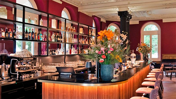 Интерьер бара Vic&rsquo;s Bar с изогнутой барной стойкой с деревянной отделкой, стеллажами с алкоголем на фоне бордовых стен и высокими стульями. / Interior of Vic&rsquo;s Bar featuring a curved bar counter with wooden trim, shelves stocked with alcohol against burgundy walls, and high stools.