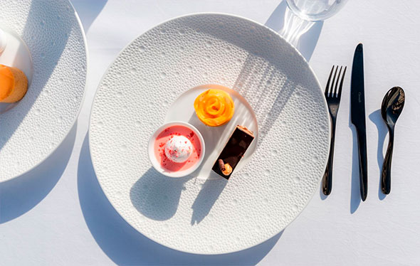 Дегустационный сет десертов (шоколадный батончик, фруктовый мусс и цитрусовый рулет) на текстурной белой тарелке, освещенной ярким солнцем.