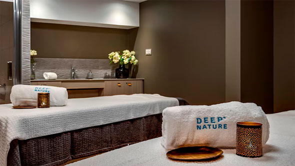 Интерьер процедурного кабинета Deep Nature Spa с двумя массажными столами, аккуратно сложенными белыми полотенцами с логотипом, цветами и свечами для расслабляющих велнес-процедур.