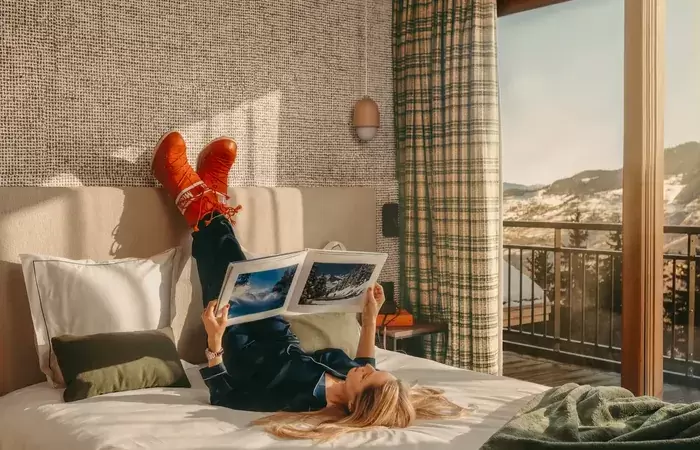 Guest reading a magazine on a bed beside a window with a mountain view, Courchevel La Tania