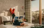 Guest reading a magazine on a bed beside a window with a mountain view, Courchevel La Tania